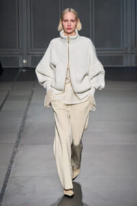 Model walking a fashion runway in an oversized white ribbed zip sweater and loose cream trousers, with a neutral expression.