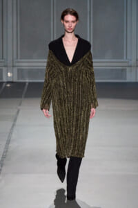 Model walking a runway in a black blazer with a deep V and a gold metallic, striped knit dress over it, with black pants visible at the bottom.