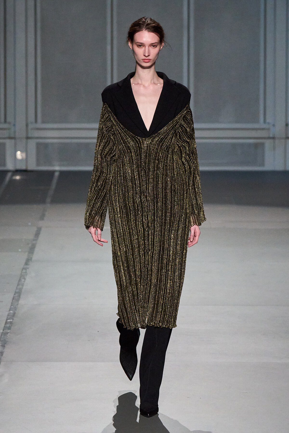 Model walking a runway in a black blazer with a deep V and a gold metallic, striped knit dress over it, with black pants visible at the bottom.