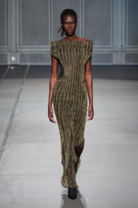 Model walks the runway in an off-the-shoulder, gold metallic striped, form-fitting floor-length dress with ribbed texture on a grey runway.
