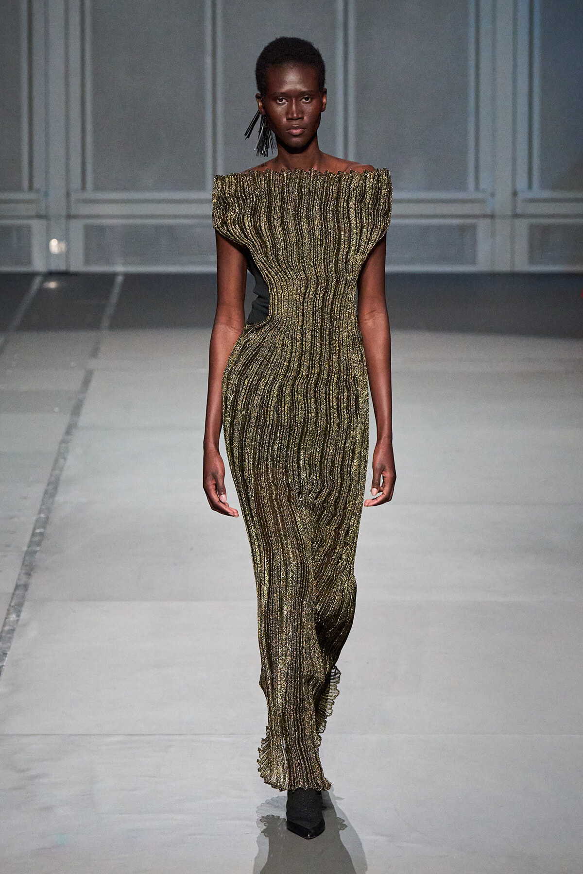 Model walks the runway in an off-the-shoulder, gold metallic striped, form-fitting floor-length dress with ribbed texture on a grey runway.