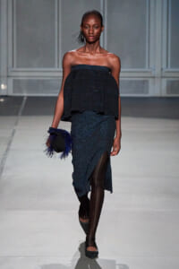 Model walks a runway in a dark blue glittery off-the-shoulder two-piece outfit with a layered top and long skirt with a thigh-high slit, carrying a black clutch with blue feathers.