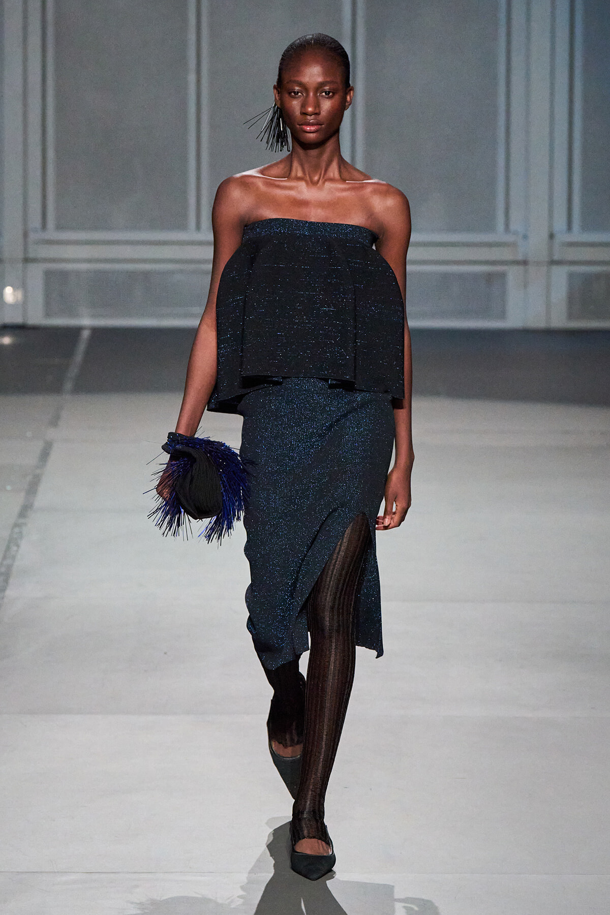 Model walks a runway in a dark blue glittery off-the-shoulder two-piece outfit with a layered top and long skirt with a thigh-high slit, carrying a black clutch with blue feathers.