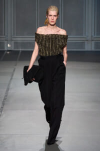 Model on a runway wearing an off‑the‑shoulder metallic striped top, black high‑waisted trousers, gold tassel earring, and a black beaded clutch.