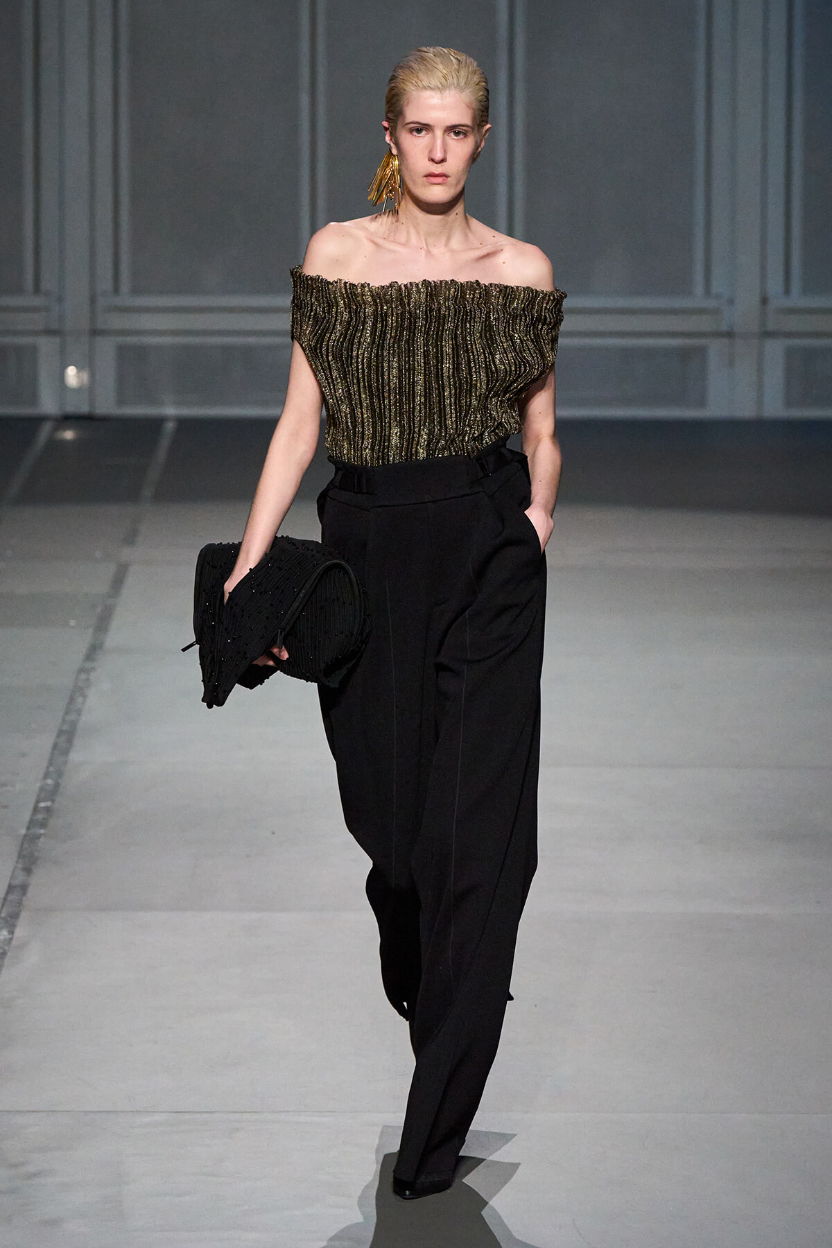 Model on a runway wearing an off‑the‑shoulder metallic striped top, black high‑waisted trousers, gold tassel earring, and a black beaded clutch.