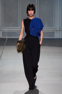 Model wearing a black sleeveless top with a bold blue draped ruffle over one shoulder, black high-waisted wide-leg pants, and a gold fringe clutch on the left hand during a runway show.