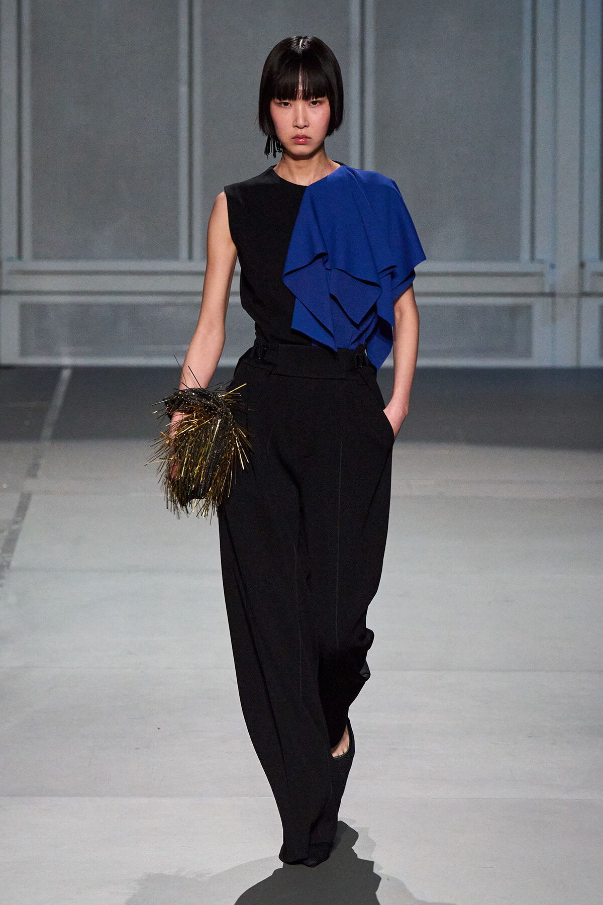 Model wearing a black sleeveless top with a bold blue draped ruffle over one shoulder, black high-waisted wide-leg pants, and a gold fringe clutch on the left hand during a runway show.