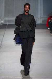 Runway model in an all-black, sculptural outfit with oversized puff sleeves and blue-thread detailing, carrying a blue spiky clutch.