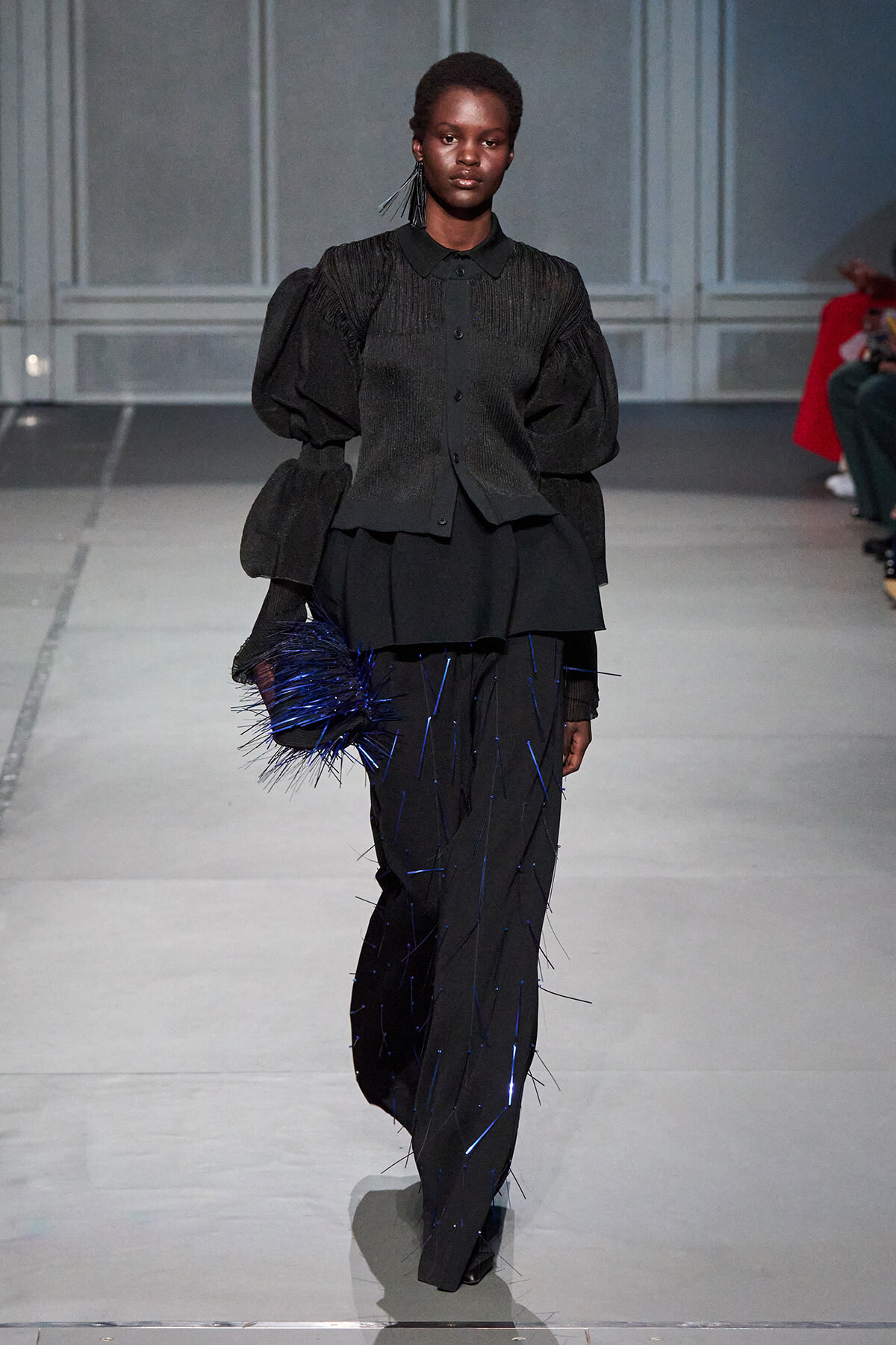 Runway model in an all-black, sculptural outfit with oversized puff sleeves and blue-thread detailing, carrying a blue spiky clutch.