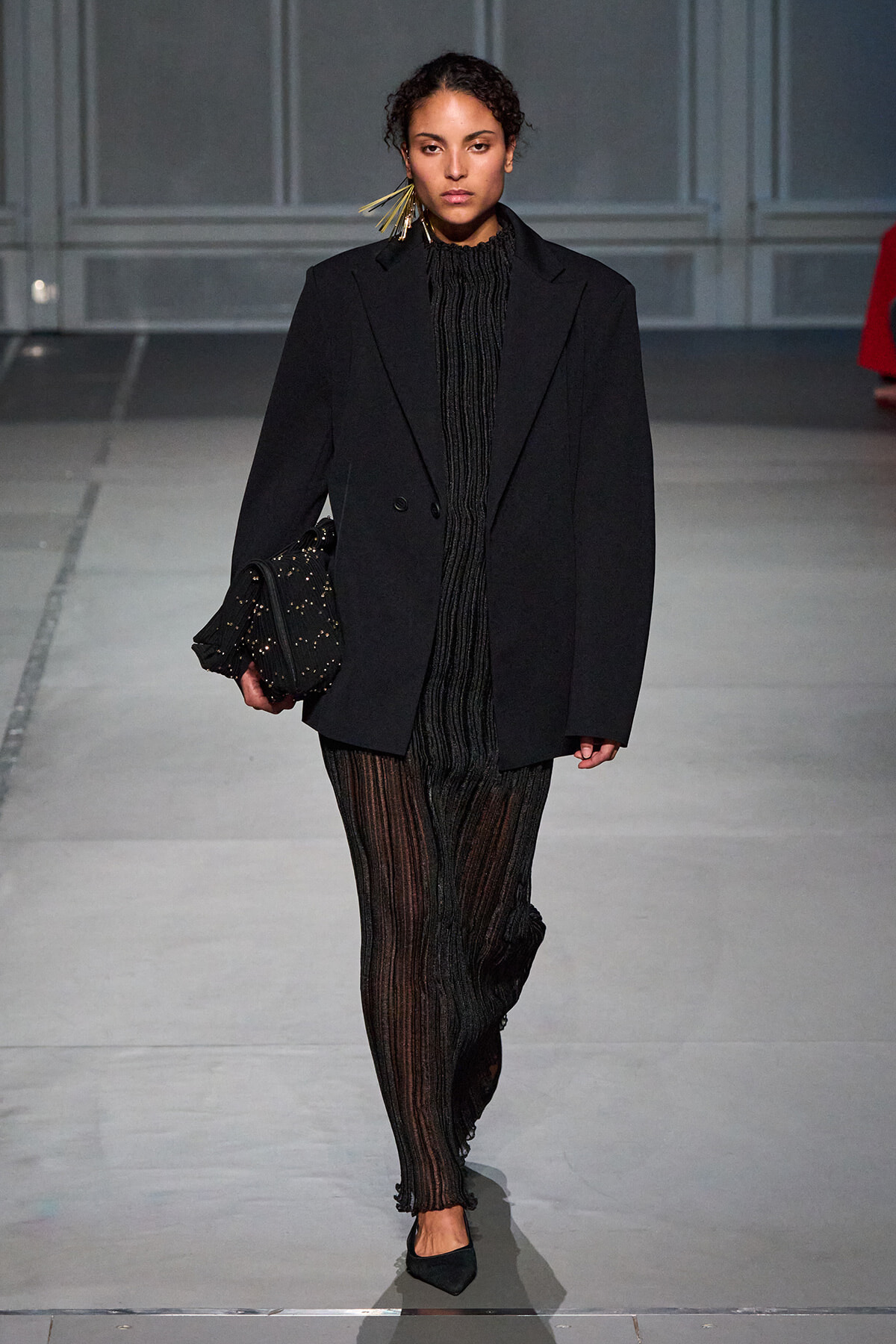 Model on a runway wearing an oversized black blazer over a sheer, striped black outfit, carrying a studded black clutch.