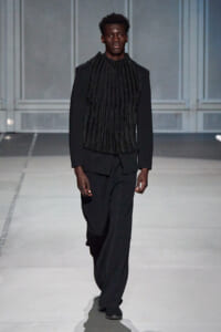 Male model walking a runway in an all-black outfit: textured ribbed scarf-like top over a blazer and wide-leg trousers.