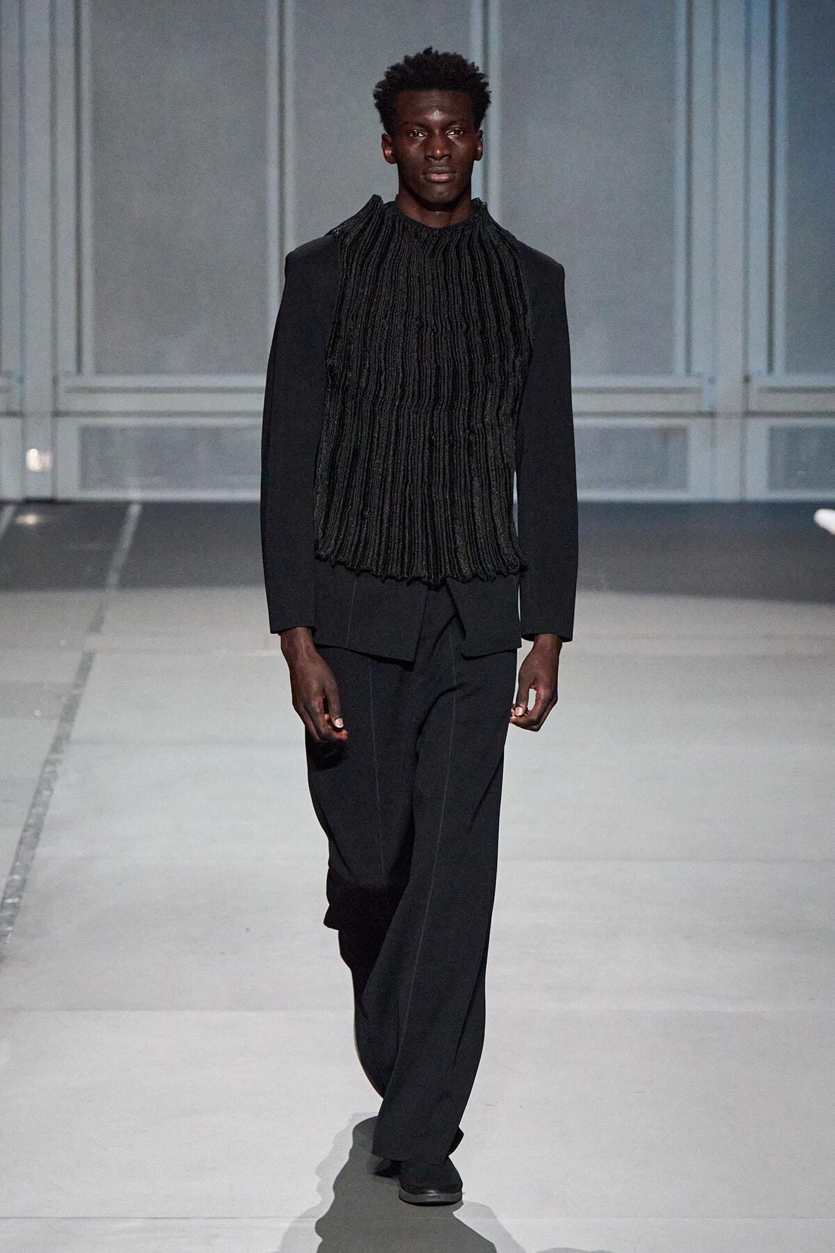 Male model walking a runway in an all-black outfit: textured ribbed scarf-like top over a blazer and wide-leg trousers.