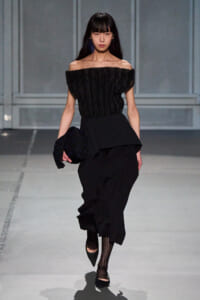 Model on a runway wearing an off-the-shoulder textured black top with a peplum waist and loose black trousers, carrying a sequined black clutch.