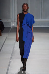 Model on runway wearing a sleeveless black dress with a bold cobalt blue draped panel over the right side, creating an asymmetric silhouette.