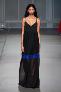 Female model walks the runway in a sleeveless black gown with a V-neck bodice and a bright blue fringe hem over a sheer skirt stretch at the bottom.