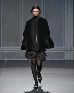 Model on a runway wearing an all-black ensemble: oversized coat over a metallic speckled dress with a fringe hem, striped tights, and black pointed flats.