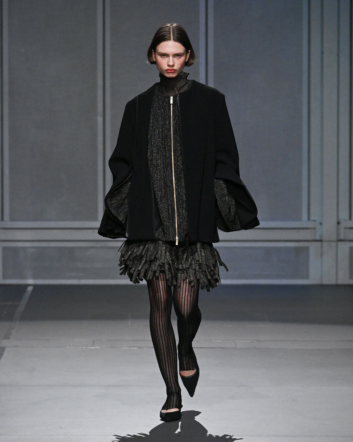 Model on a runway wearing an all-black ensemble: oversized coat over a metallic speckled dress with a fringe hem, striped tights, and black pointed flats.