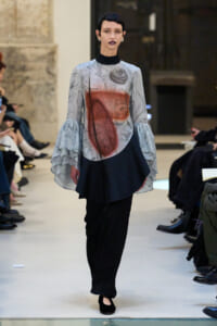 Model walking a runway in a marbled gray top with dramatic ruffled sleeves and black wide-leg pants.