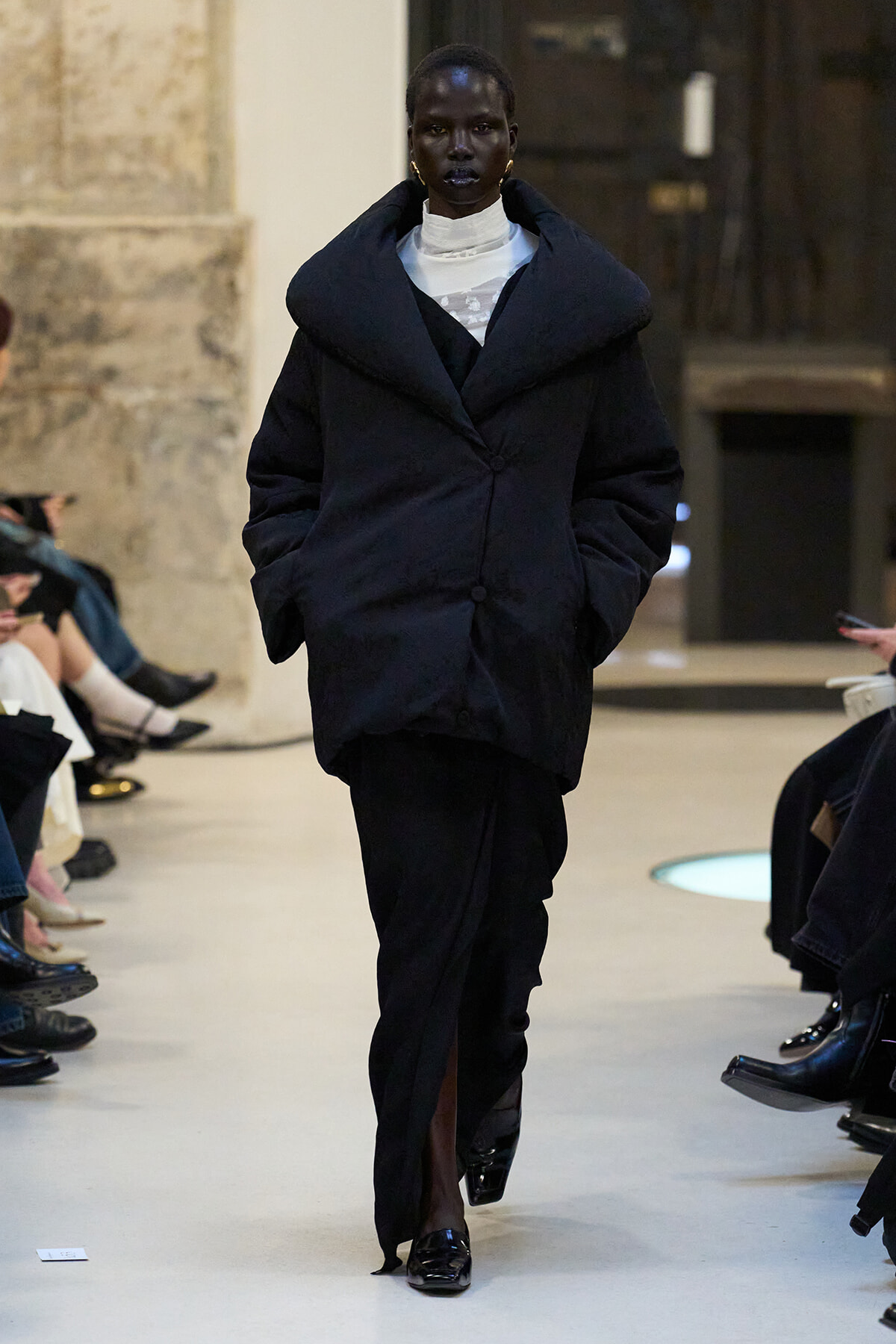 Model wearing an oversized black coat with a wide collar, walking the runway beside seated audience members.