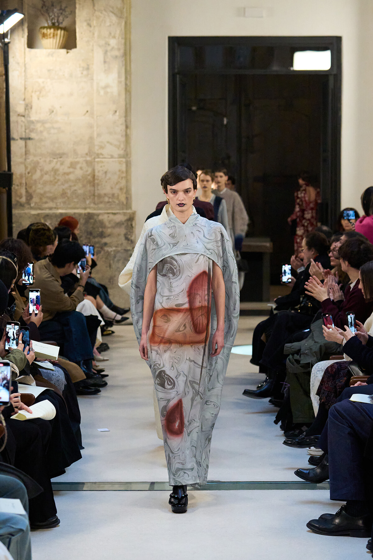 Model walking a marble-patterned silver gown with red shapes down a runway, audience filming on both sides.