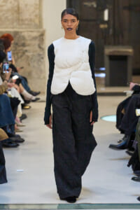 Model walking the runway in a white sculptural padded vest over a black long-sleeve top and wide-leg black pants, with audience along the sides.
