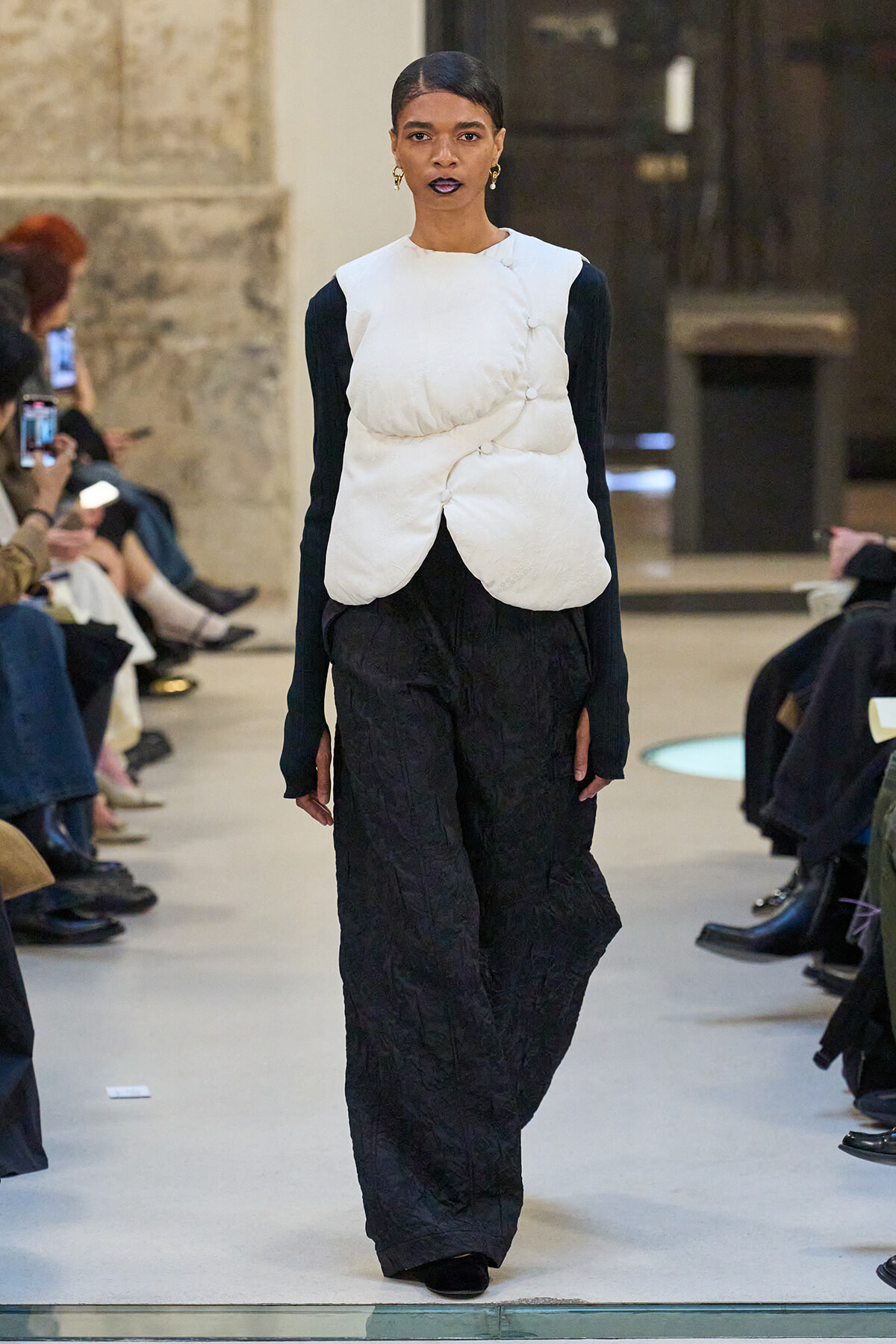 Model walking the runway in a white sculptural padded vest over a black long-sleeve top and wide-leg black pants, with audience along the sides.