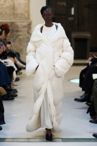 Model walks down runway in an oversized white padded coat with a draped scarf-like collar, black lipstick, and braided hair.