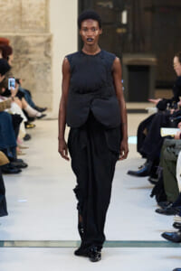 Model walks a runway in an all-black outfit: sleeveless draped top with wrap detailing and wide, flowing black pants; glossy black shoes.