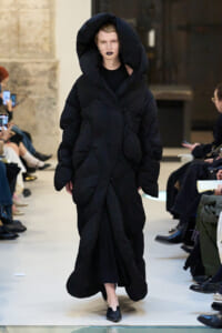Model walking a runway in an oversized black puffer coat with a large hood and dark lipstick.