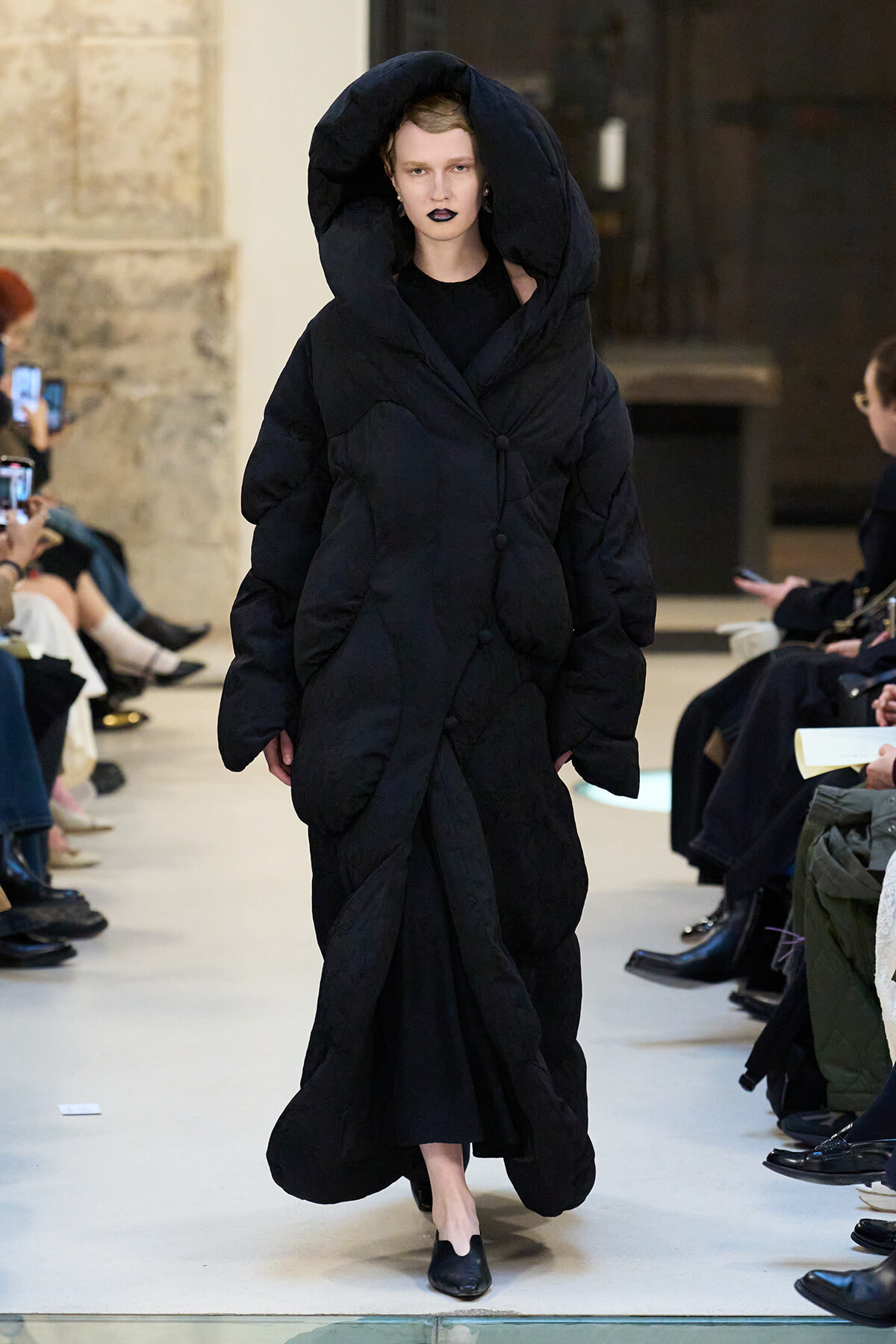 Model walking a runway in an oversized black puffer coat with a large hood and dark lipstick.