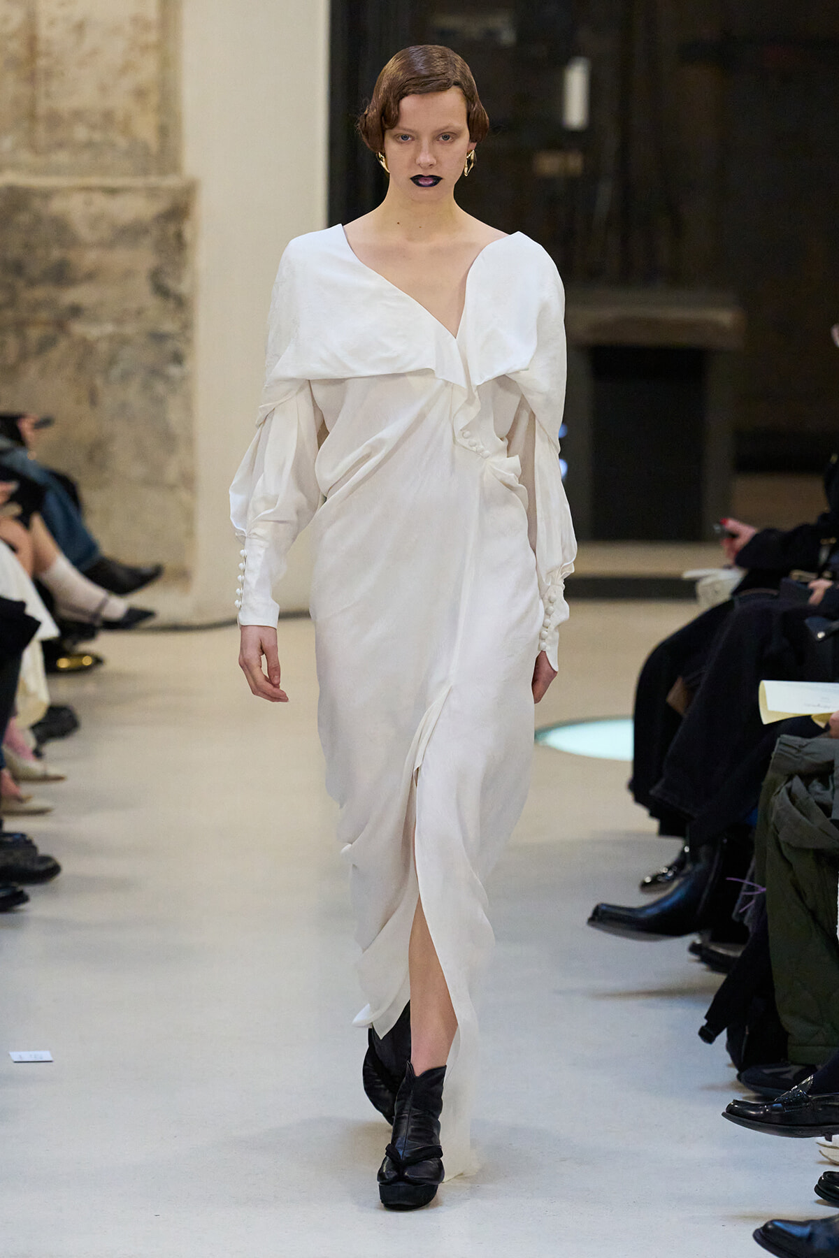 Model walking a runway in a white draped wrap dress with an off-shoulder neckline and black ankle boots.