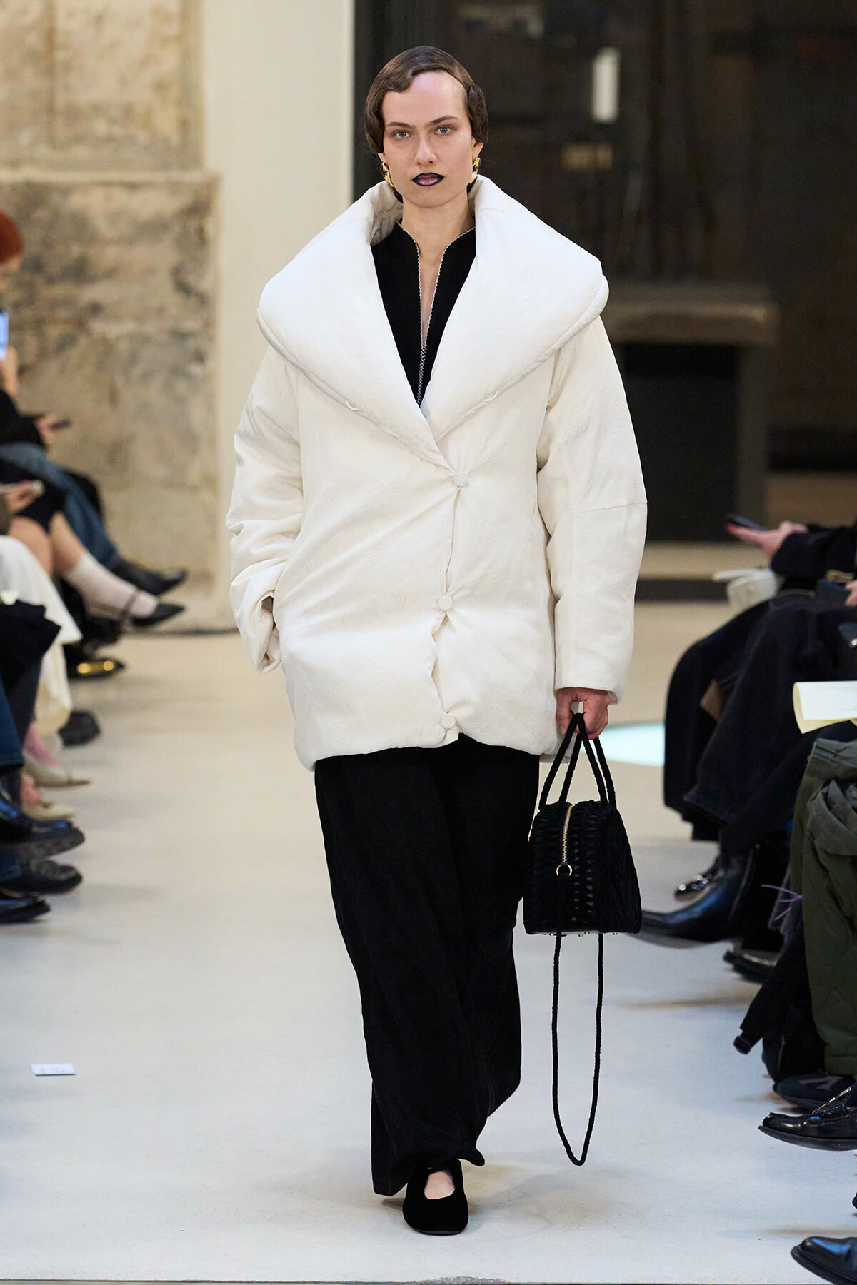 Model walks a runway in a voluminous white coat with oversized collar, black outfit, and black handbag.