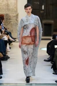 Model walks the runway in a flowing grey marble-pattern cape dress with red abstract patches at the torso, wearing dark lipstick.