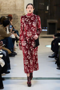 Model walks the runway in a burgundy velvet, high-neck dress with a white ornate pattern and a black shoulder bag.