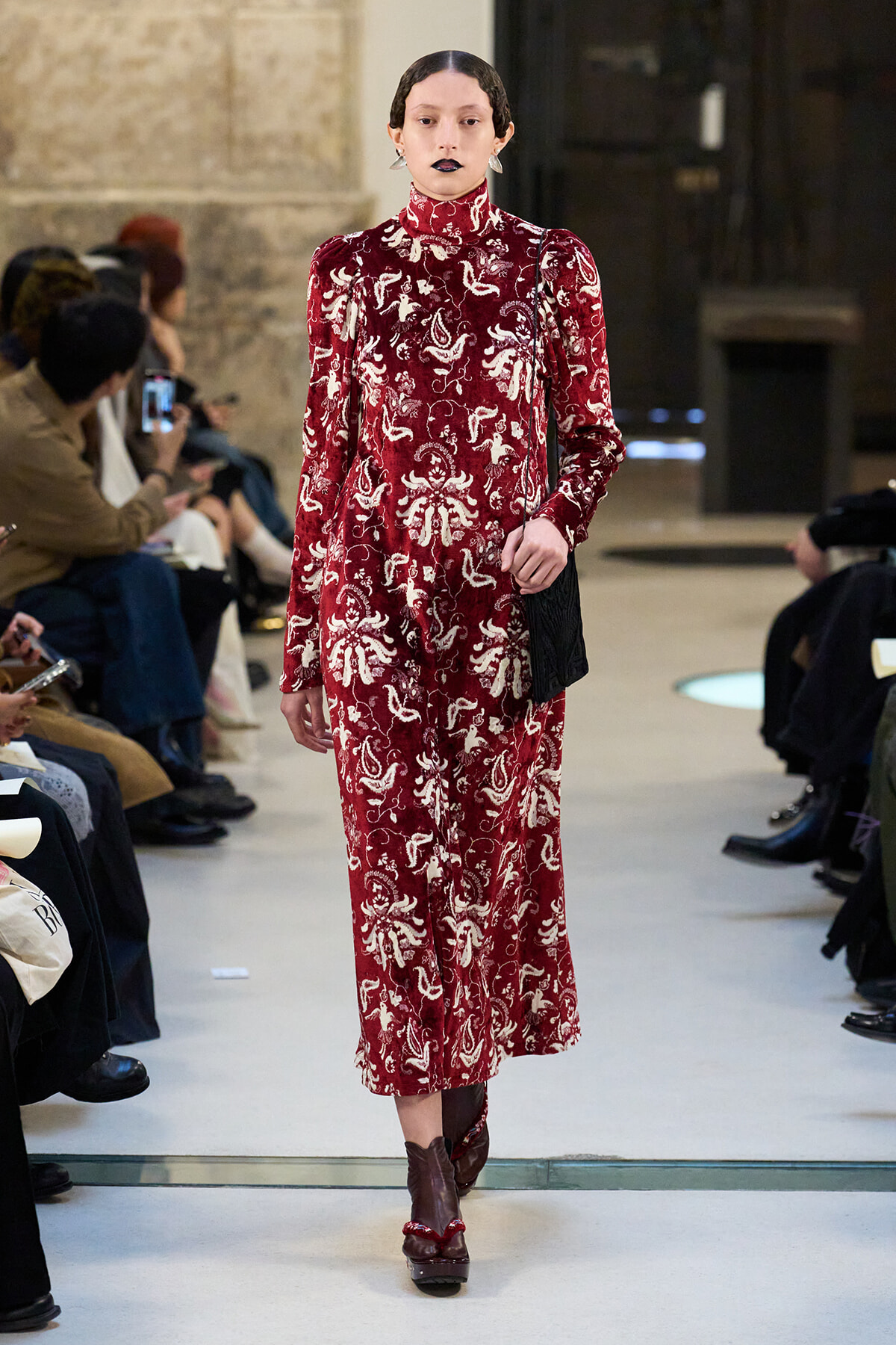 Model walks the runway in a burgundy velvet, high-neck dress with a white ornate pattern and a black shoulder bag.