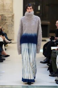 Model walks a runway in a fluffy gray turtleneck sweater and a blue-to-white striped pleated skirt with a dark blue hem at the bottom.