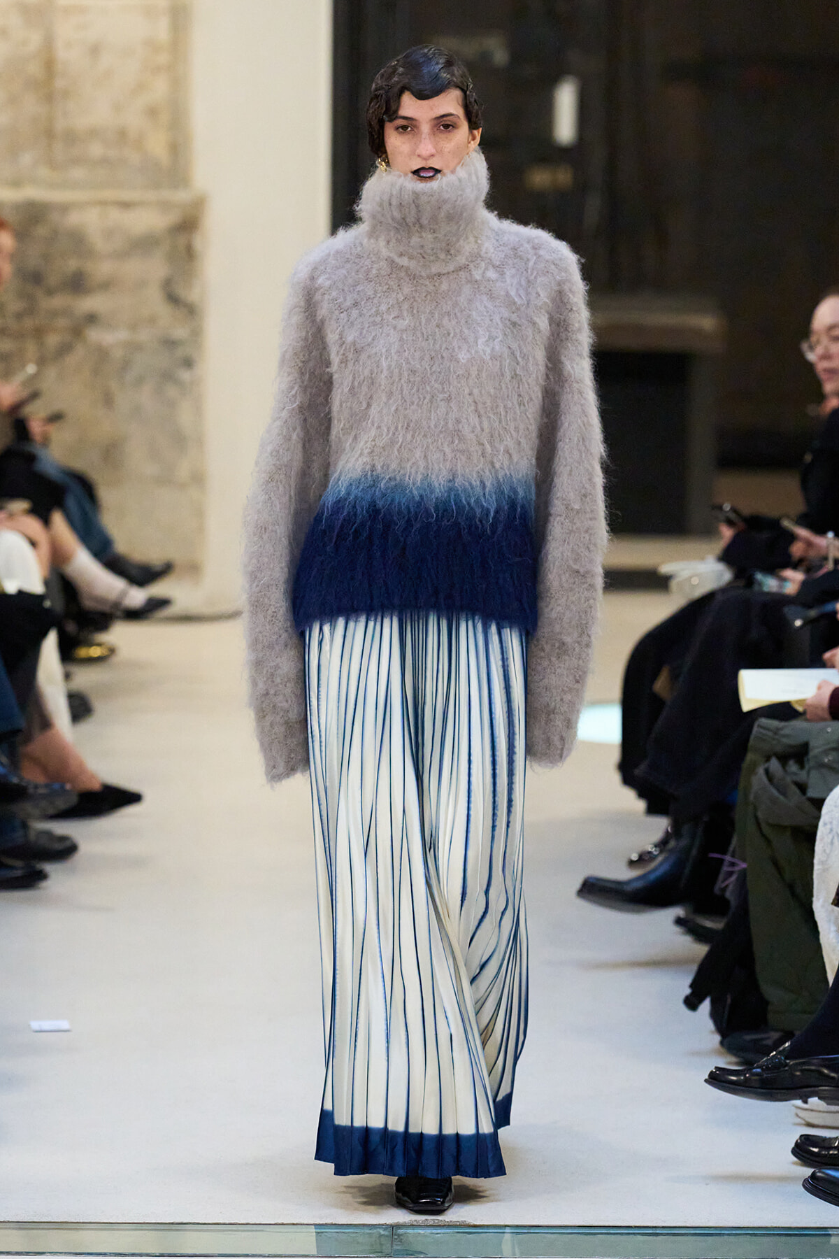 Model walks a runway in a fluffy gray turtleneck sweater and a blue-to-white striped pleated skirt with a dark blue hem at the bottom.