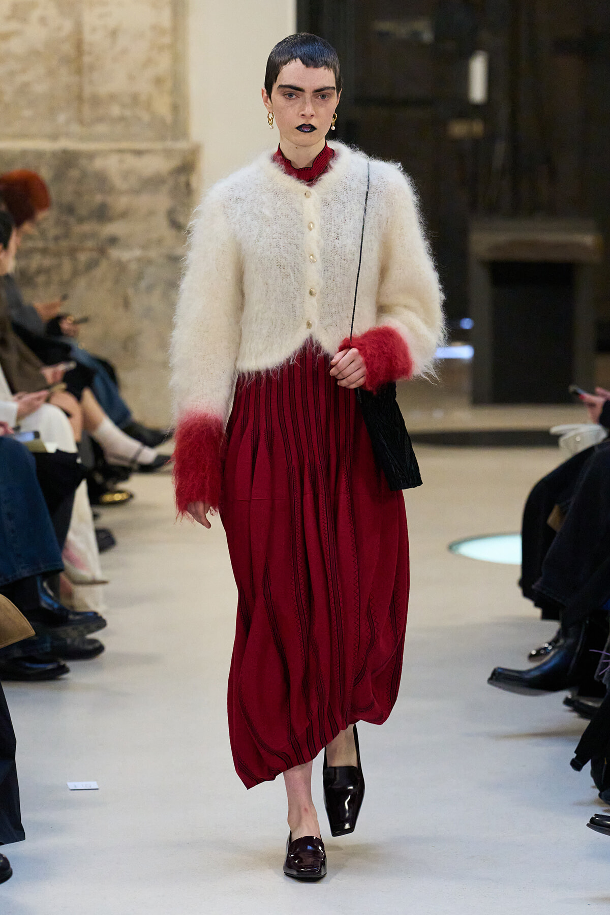 Model walking the runway in an ivory fuzzy cardigan over a red pleated midi dress with red cuffs, carrying a black shoulder bag.