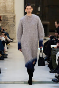 Model walks a runway in a oversized fuzzy gray knit dress with dark blue cuffs and hem, carrying a black handbag.