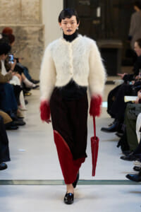 Model walking a runway in a fluffy white cardigan with red-dyed cuffs, black turtleneck, and red-and-black patchwork pants, carrying a red bag.
