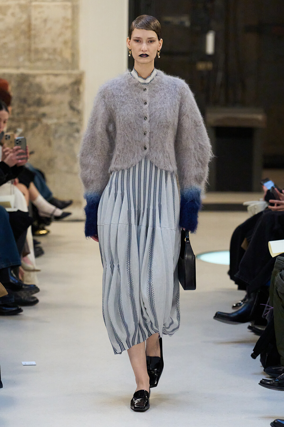 Model walking a runway in a light gray fuzzy cardigan over a blue striped midi dress, carrying a small black bag.