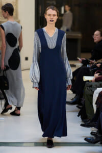 Model on a runway wearing a navy blue sleeveless dress over a striped blouse with voluminous sleeves.