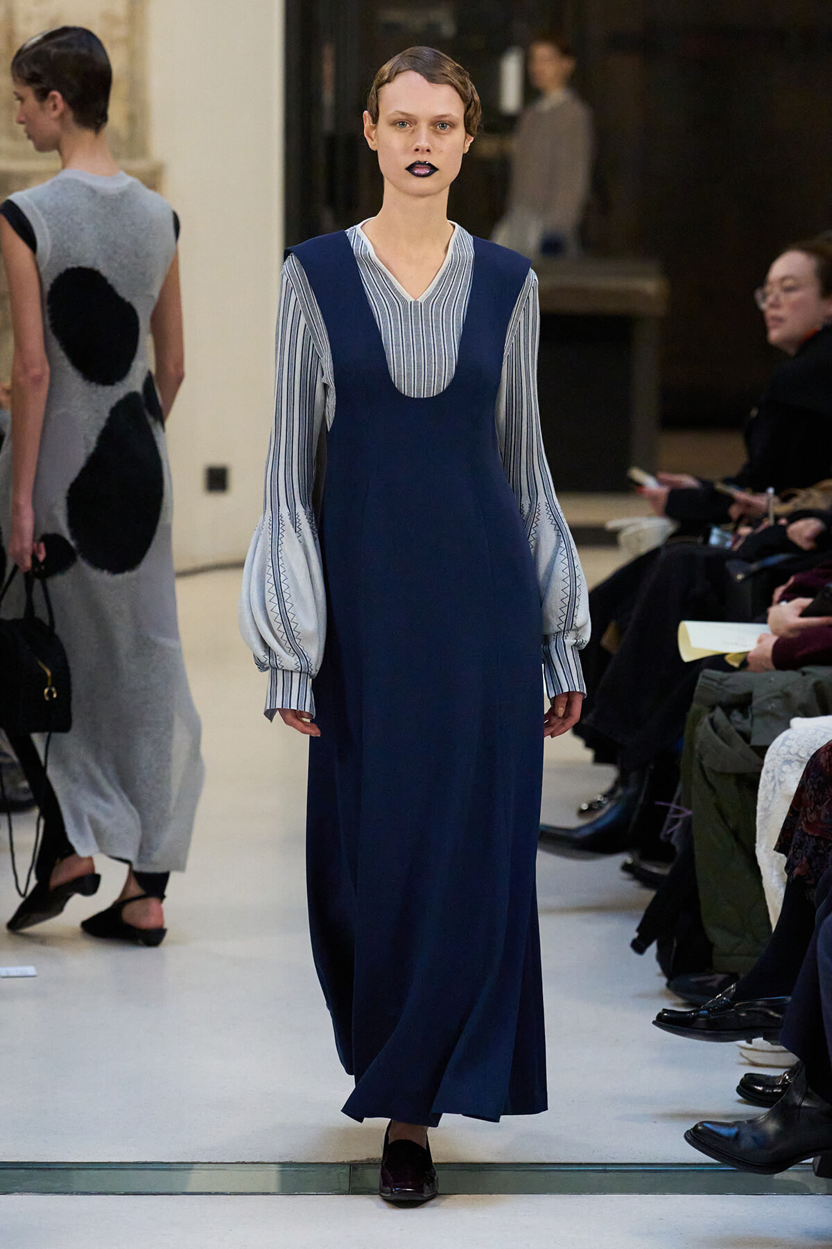Model on a runway wearing a navy blue sleeveless dress over a striped blouse with voluminous sleeves.