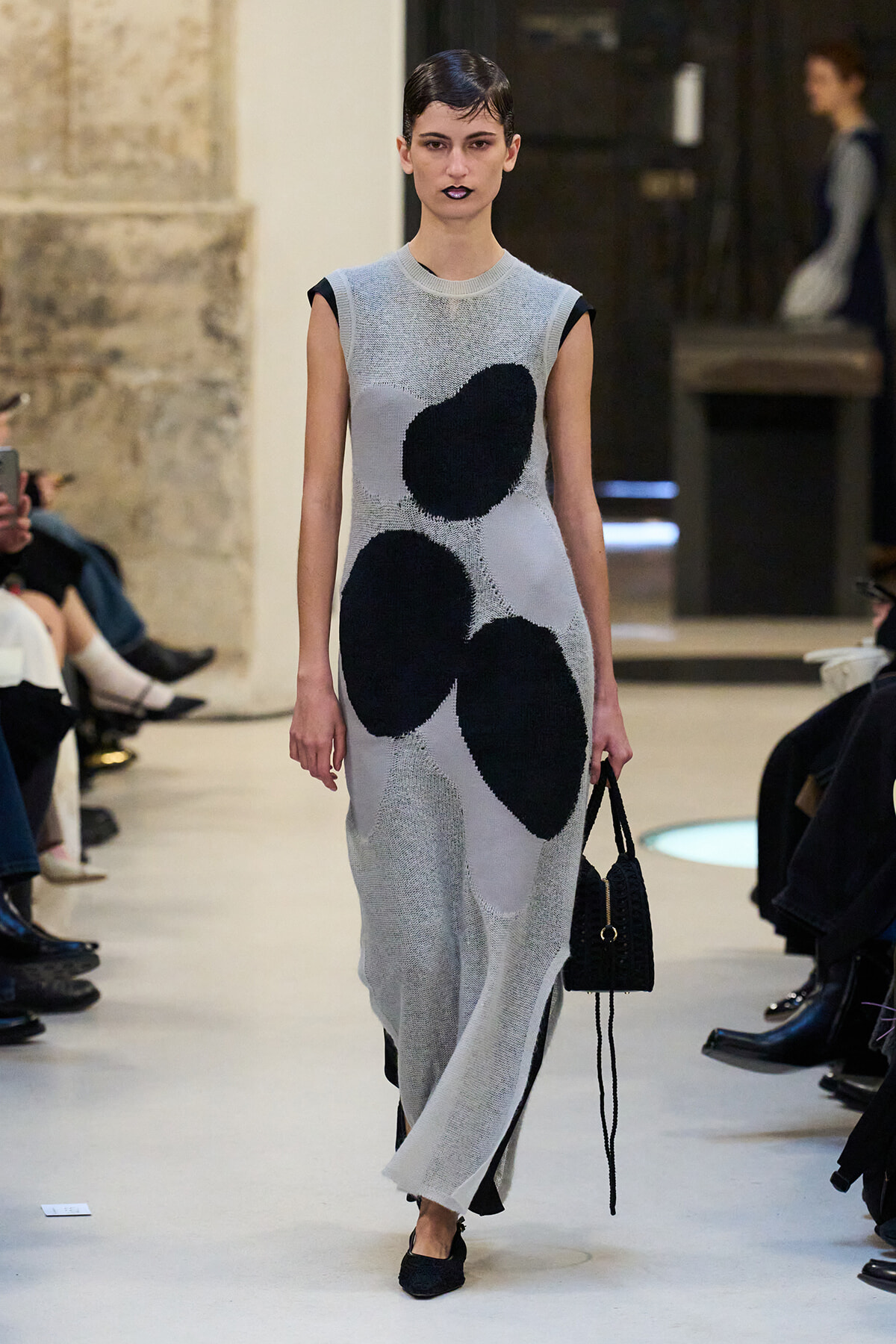 Model on a runway wearing a sleeveless light-gray knit maxi dress with large black abstract patches, carrying a small black handbag.