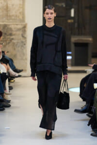 Model walking a runway in an all-black knit set with a long skirt, carrying a black woven handbag.