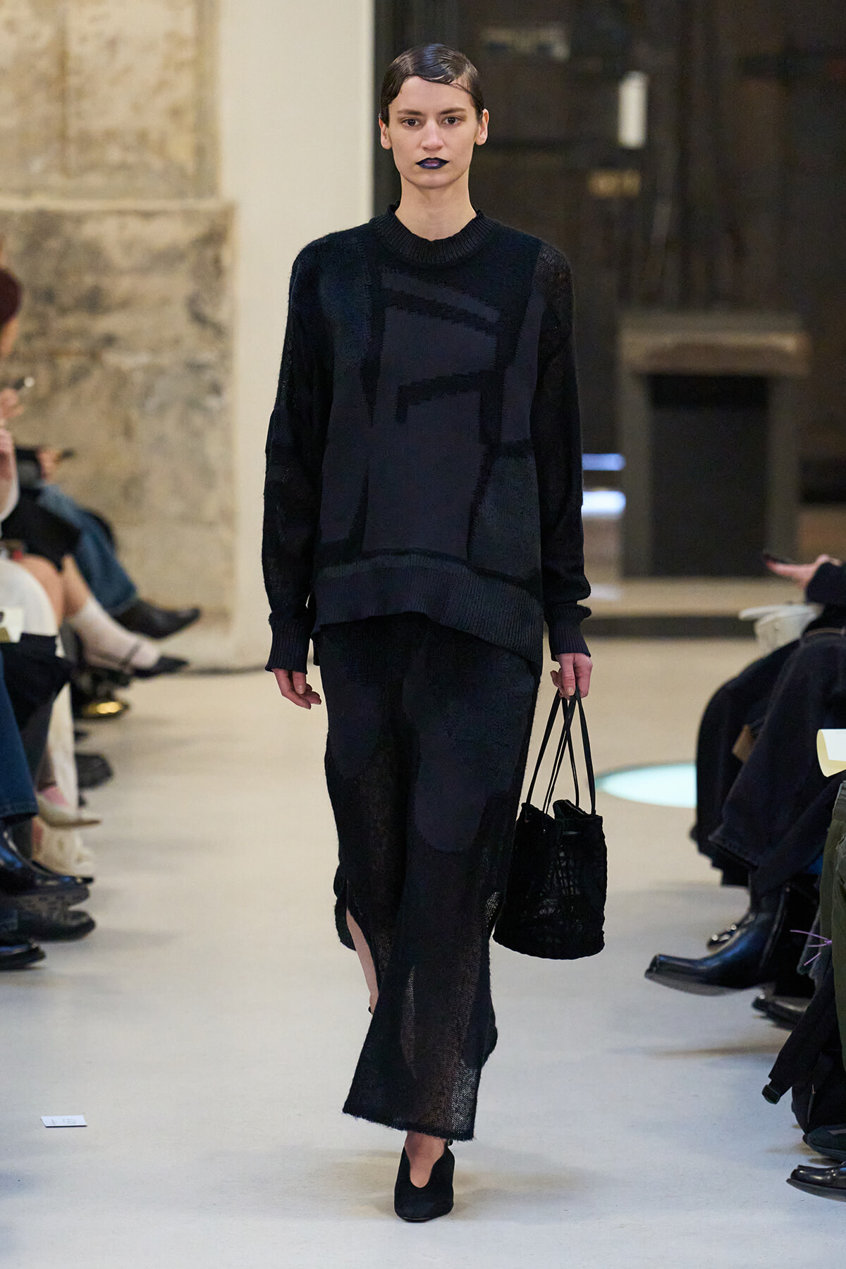 Model walking a runway in an all-black knit set with a long skirt, carrying a black woven handbag.