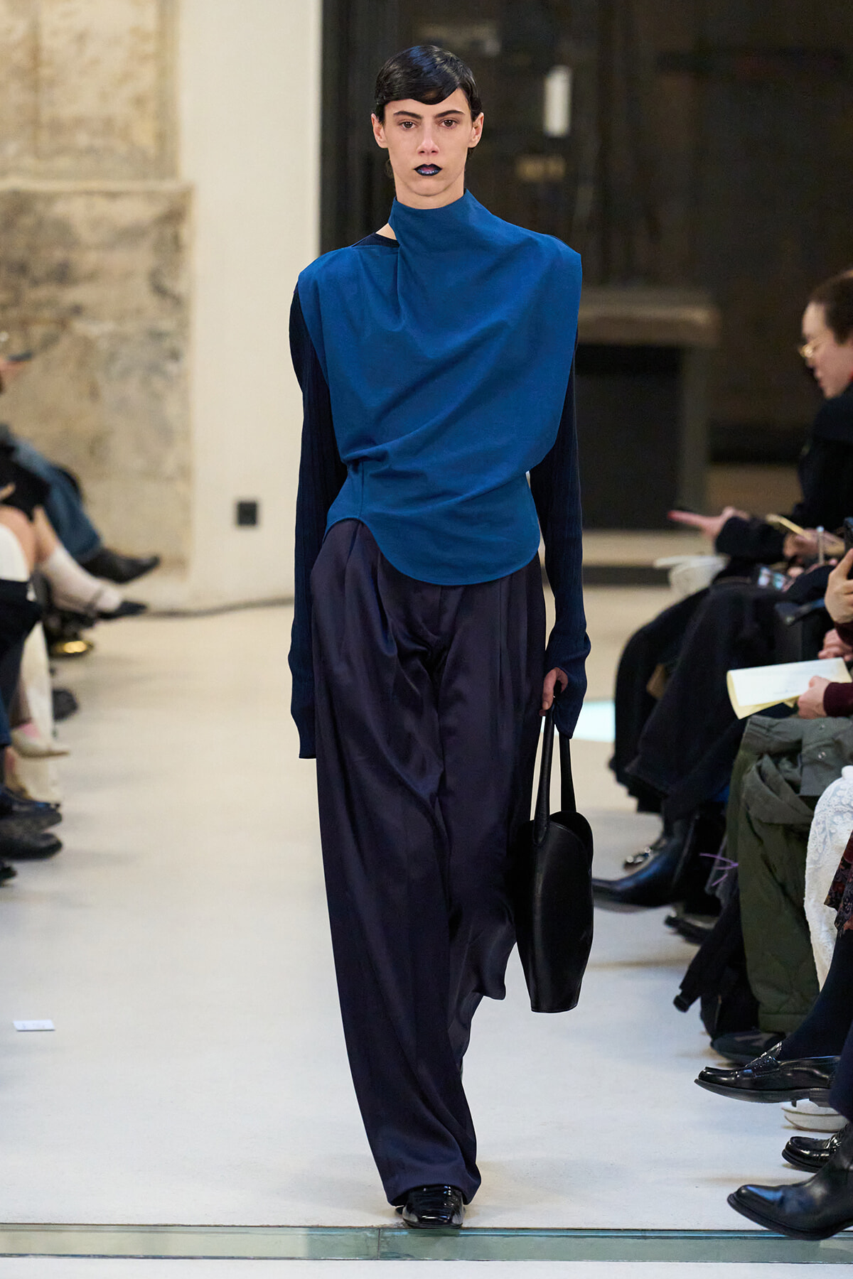 Model walks the runway in a cobalt blue draped top and loose navy trousers, carrying a black handbag.