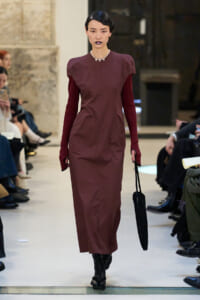 Runway model in a burgundy midi dress with puff shoulders, pearl necklace, dark lipstick, black knit handbag, and boots.