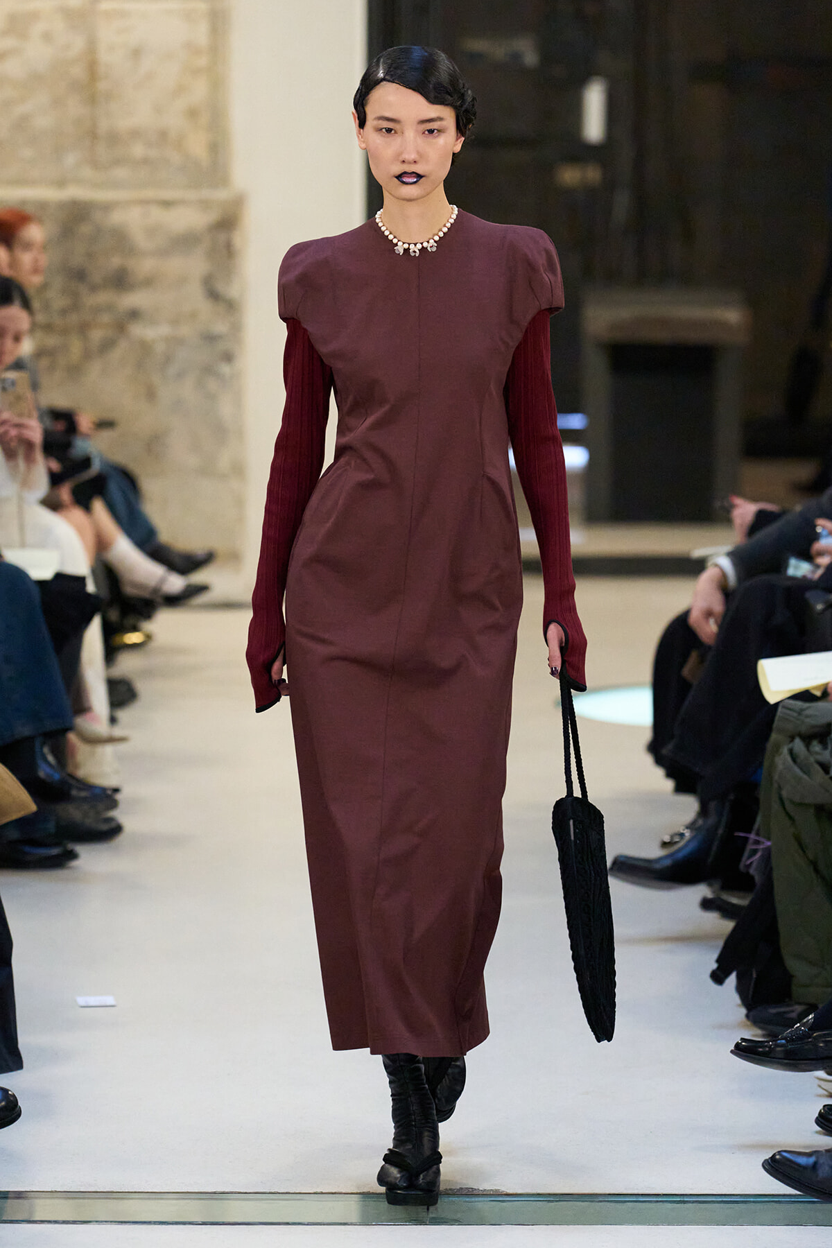 Runway model in a burgundy midi dress with puff shoulders, pearl necklace, dark lipstick, black knit handbag, and boots.