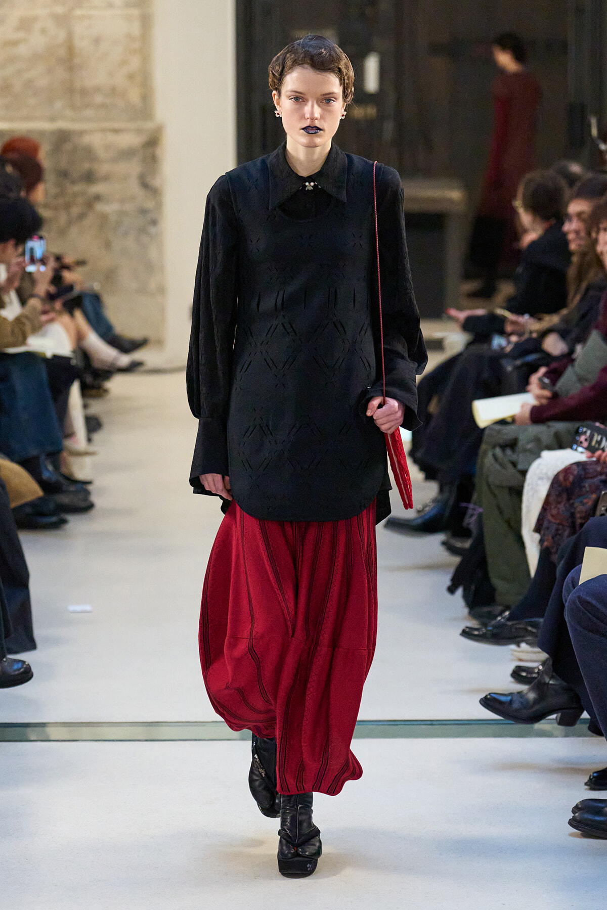 Model walks a fashion show wearing a black textured blouse and a voluminous red skirt, holding a red bag, with an audience along the runway behind her.